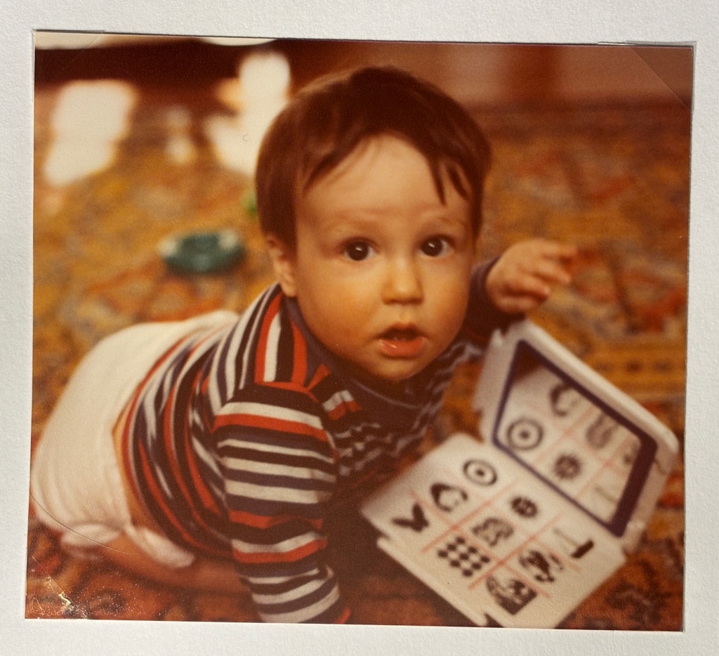 Jacob Lodwick as a baby, crawling on a carpet holding a flip book of symbols